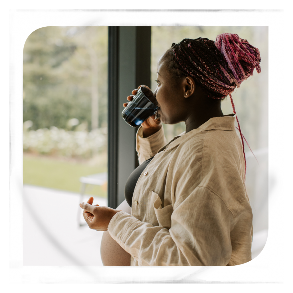A young pregnant woman drinking water while she stares out of her glass doors.