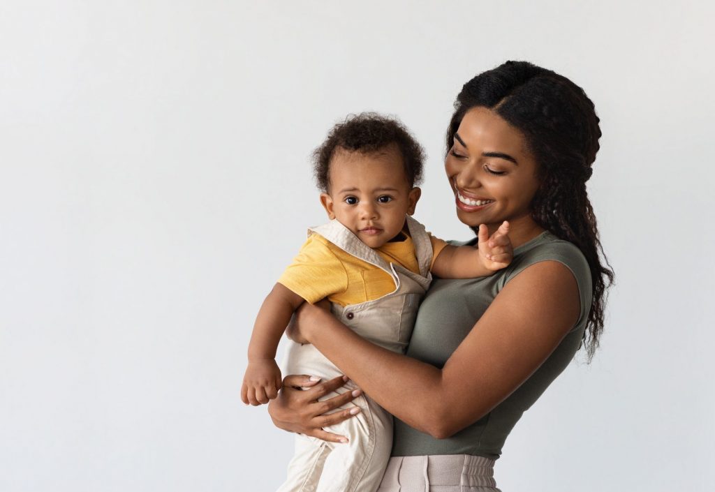 Portrait Of Smiling Young Black Mother Holding Adorable Infant Son On Arms.